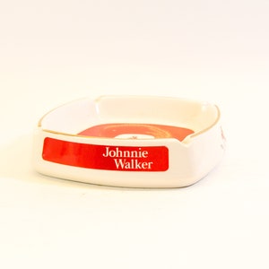 May include: White ceramic ashtray with a red and gold design. The ashtray has a rectangular shape with rounded corners and a gold trim. The words "Johnnie Walker" are printed on a red label.
