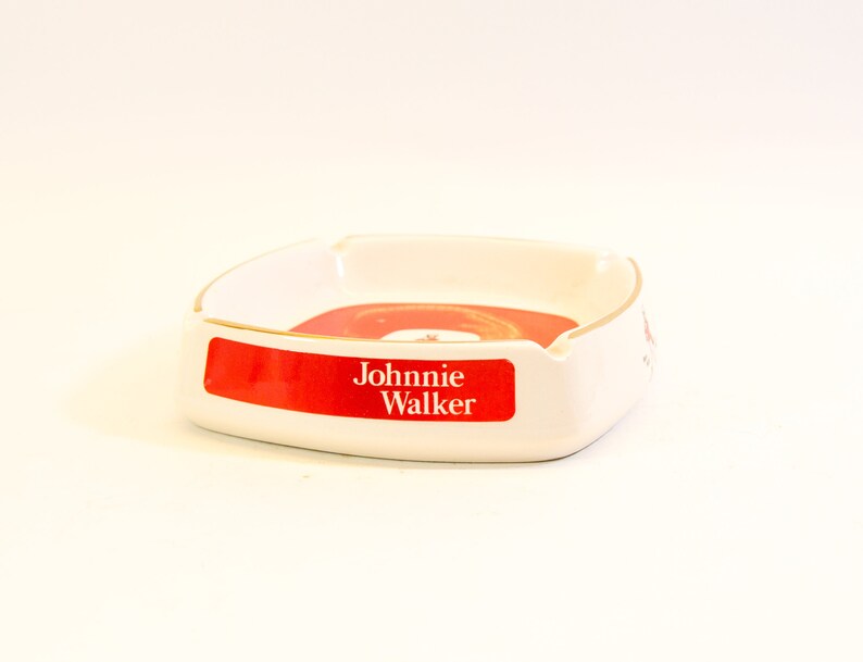 May include: White ceramic ashtray with a red and gold design. The ashtray has a rectangular shape with rounded corners and a gold trim. The words "Johnnie Walker" are printed on a red label.