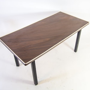 Op de afbeelding: Een rechthoekige salontafel met een donkerbruin houten blad en een zilveren rand. De tafel heeft zwarte, vierkante poten. De tafel is ongeveer 122 cm lang en 61 cm breed.