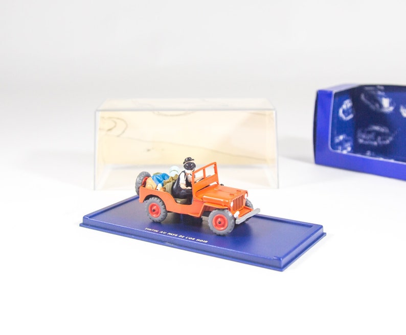 May include: A red toy jeep with a black driver figure and a blue and white box. The jeep has a black roof and black wheels. The box has the words 'The Toys of Our Time' printed on the bottom.