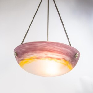 Pu&ograve; includere: Una lampada a sospensione con un paralume a forma di ciotola. Il paralume presenta una miscela di colori, tra cui rosa, viola, giallo e bianco, creando un effetto tramonto. La luce &egrave; sospesa da tre fili metallici.