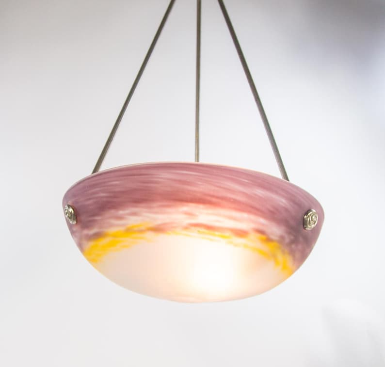 Pu&ograve; includere: Una lampada a sospensione con un paralume a forma di ciotola. Il paralume presenta una miscela di colori, tra cui rosa, viola, giallo e bianco, creando un effetto tramonto. La luce &egrave; sospesa da tre fili metallici.