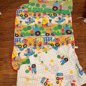 以下が含まれることがあります： セサミストリートの寝具セット。キルトブランケット1枚と枕カバー2枚が含まれます。生地には、エルモ、ビッグバード、クッキーモンスターなどのカラフルなキャラクターが、トラクター、列車、飛行機などの様々な乗り物を操縦している様子が描かれています。