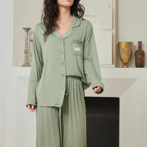 Puede incluir: Conjunto de pijama verde salvia con mangas largas y pantalón. La parte superior tiene una botonadura y un bolsillo en el pecho con las iniciales "MK". El pantalón tiene cintura con cordón. El conjunto es de tejido suave y fluido.