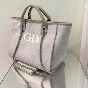 May include: A beige canvas tote bag with a silver chain strap and the initials "GD" embroidered on the front.
