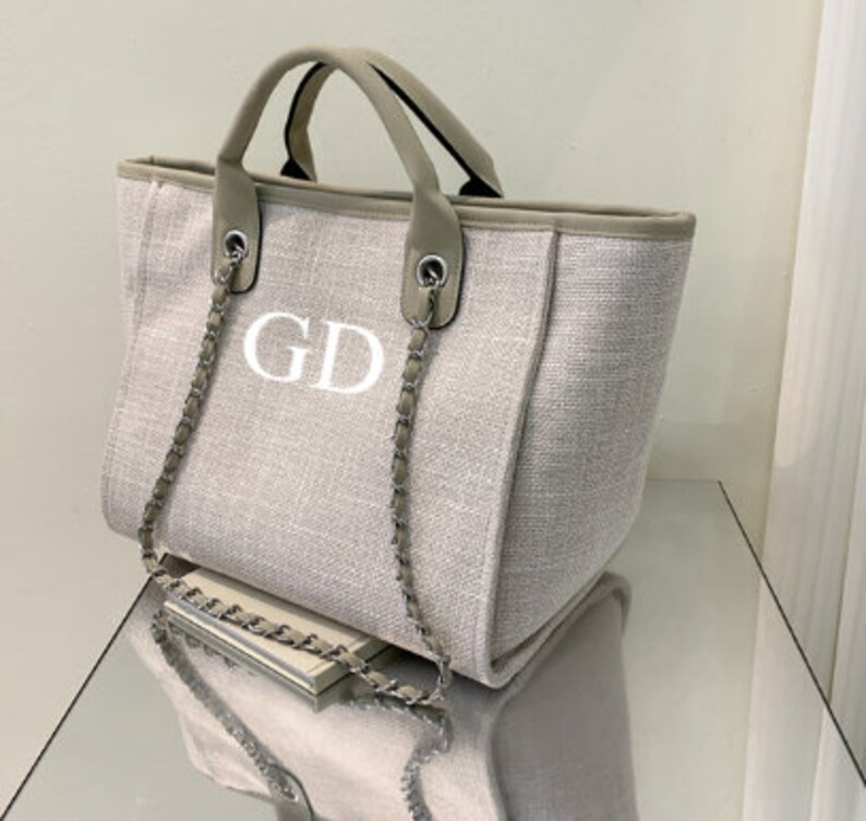 May include: A beige canvas tote bag with a silver chain strap and the initials "GD" embroidered on the front.