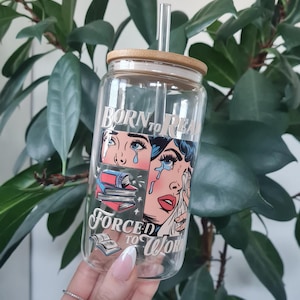 May include: A clear glass water bottle with a bamboo lid and a straw. The bottle features a comic book style graphic of a woman crying with the text "Born to Read, Forced to Work".