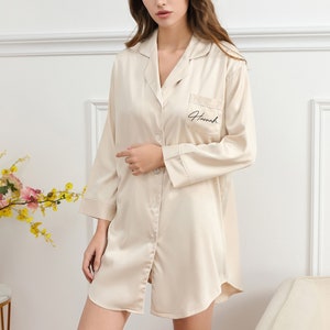 May include: A beige satin sleep shirt with long sleeves and a button-up front. The shirt has a patch pocket on the left chest with the name "Hannah" embroidered in black.