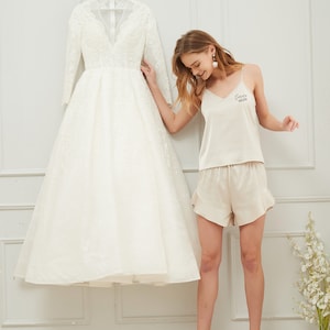 May include: A woman in a white satin pajama top and matching shorts with the text "Future Bride" printed on the top. She is standing in front of a white wall with a white wedding dress hanging on a hanger.