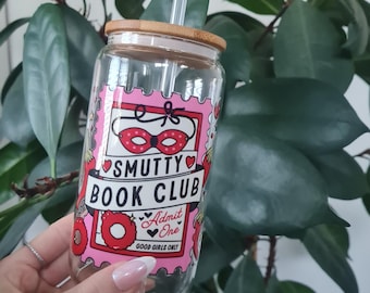 May include: A clear glass reusable water bottle with a bamboo lid and a straw. The bottle has a pink and red design with the text "Smutty Book Club" and "Good Girls Only".