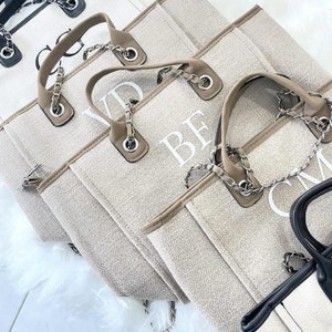May include: Four beige canvas tote bags with chain straps and white embroidered letters. The bags are lined up in a row, with the letters "VD", "BF", "CM" and a fourth bag with a black chain strap visible.
