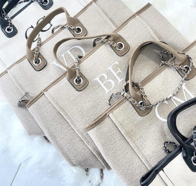 May include: Four beige canvas tote bags with chain straps and white embroidered letters. The bags are lined up in a row, with the letters "VD", "BF", "CM" and a fourth bag with a black chain strap visible.