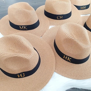 May include: Five straw hats with black bands and gold initials. The initials are MK, CH, MH, HK, and HJ.