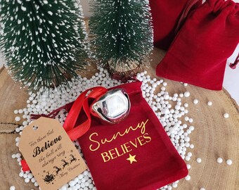May include: A silver jingle bell with a red ribbon sits on a red velvet drawstring pouch printed with the gold text Sunny BELIEVES and a star. A tan gift tag reads For those that Believe the bell will always ring Merry Christmas.