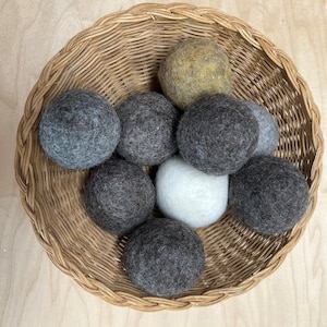 Peut inclure: Un panier rond en osier tressé rempli de plusieurs boules de séchage en laine feutrée. Les boules sont de différentes nuances de gris, blanc et vert olive. Le panier est de couleur marron clair naturelle.