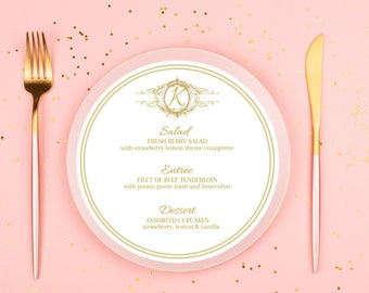 May include: A white plate with a gold border and a gold letter K monogram in the center. The plate has a menu printed on it with the headings Salad, Entree, and Dessert. The menu items are: Fresh Berry Salad with strawberry lemon thyme vinaigrette, Filet of Beef Tenderloin with potato puree hash and broccolini, and Assorted Cupcakes strawberry, lemon & vanilla.