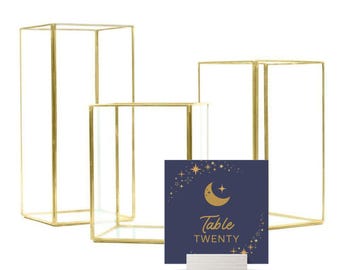 May include: Three gold metal geometric glass vases of different sizes with a navy blue card with gold stars and a crescent moon and the text "Table Twenty" on a white stand.