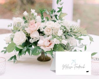 May include: White table with a white and pink floral centerpiece. A white card with the text "Melissa Frederick" and a blue ribbon is on the table.