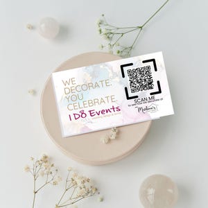 Puede incluir: Una tarjeta de visita blanca con un fondo de acuarela y texto dorado. El texto dice "We decorate. You celebrate. I DO Events wedding design & rentals". Un código QR se encuentra en la esquina superior derecha con el texto "Scan me to see how we decorate at Marlene's Event Design".