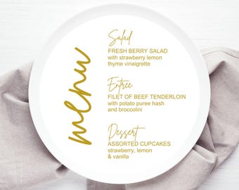 May include: A white plate with a gold menu written on it. The menu includes a salad, entree, and dessert. The salad is fresh berry salad with strawberry lemon thyme vinaigrette. The entree is filet of beef tenderloin with potato puree hash and broccolini. The dessert is assorted cupcakes, strawberry, lemon, and vanilla.
