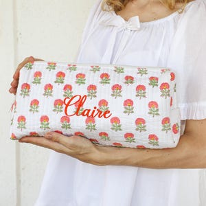 May include: A rectangular white fabric pouch featuring a repeating pattern of small pink and orange flowers with green stems. The name Claire is embroidered in red cursive script across the center of the front panel of the zippered bag.