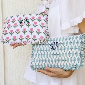 Puede incluir: Dos clutches rectangulares decorativos. Uno presenta un fondo blanco con un estampado floral rosa e iniciales verdes. El otro tiene un diseño geométrico azul y blanco con iniciales azules. Ambos tienen bordes con volantes.