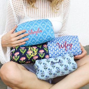 May include: A collection of cosmetic bags in various colors and patterns. The bags feature embroidered names and monograms. Designs include floral, geometric, and paisley patterns. Colors include blue, navy, and light blue.