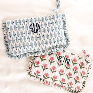 May include: Two quilted cosmetic bags. One is white with a blue floral pattern and the initials "SLA" embroidered in navy. The other is white with a pink floral pattern and the initials "K/N" embroidered in green. Both have ruffled edges.