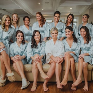 May include: A group of women wearing light blue satin robes, with one woman in the center wearing a white satin robe. They are all sitting on a couch and smiling at the camera.