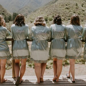 May include: Six pale green satin robes with lace trim, each embroidered with a different name in cursive. The women are standing on a wooden deck overlooking a mountain range. The robes are tied at the waist.