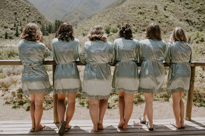 May include: Six pale green satin robes with lace trim, each embroidered with a different name in cursive. The women are standing on a wooden deck overlooking a mountain range. The robes are tied at the waist.