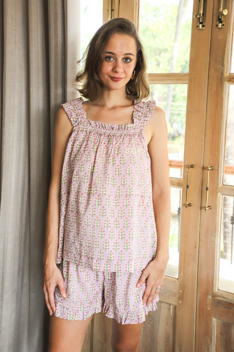 May include: A light purple sleeveless top and matching shorts set featuring a repeating floral pattern of small pink flowers with green stems. The top has a square neckline with ruffled shoulder straps, and the shorts feature a ruffled hemline.