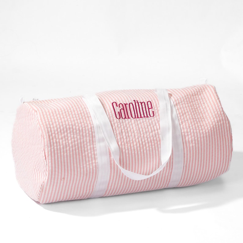Puede incluir: Bolsa de viaje de lona a rayas rosa y blancas con asas blancas y el nombre "Caroline" bordado en rojo.