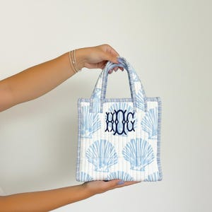May include: A small, quilted tote bag with a white background and a pattern of light blue seashells. The bag has a navy blue monogram and trim. The handles are also light blue and white striped. The bag is held by a person.