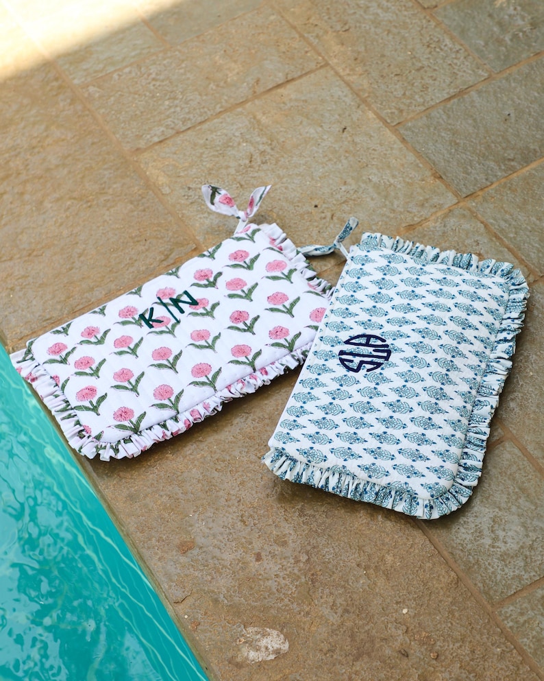May include: Two rectangular decorative cushions with floral and patterned designs. One cushion features pink flowers on a white background with the initials "K/N" embroidered in green. The other has a blue pattern on a white background with a blue monogram.