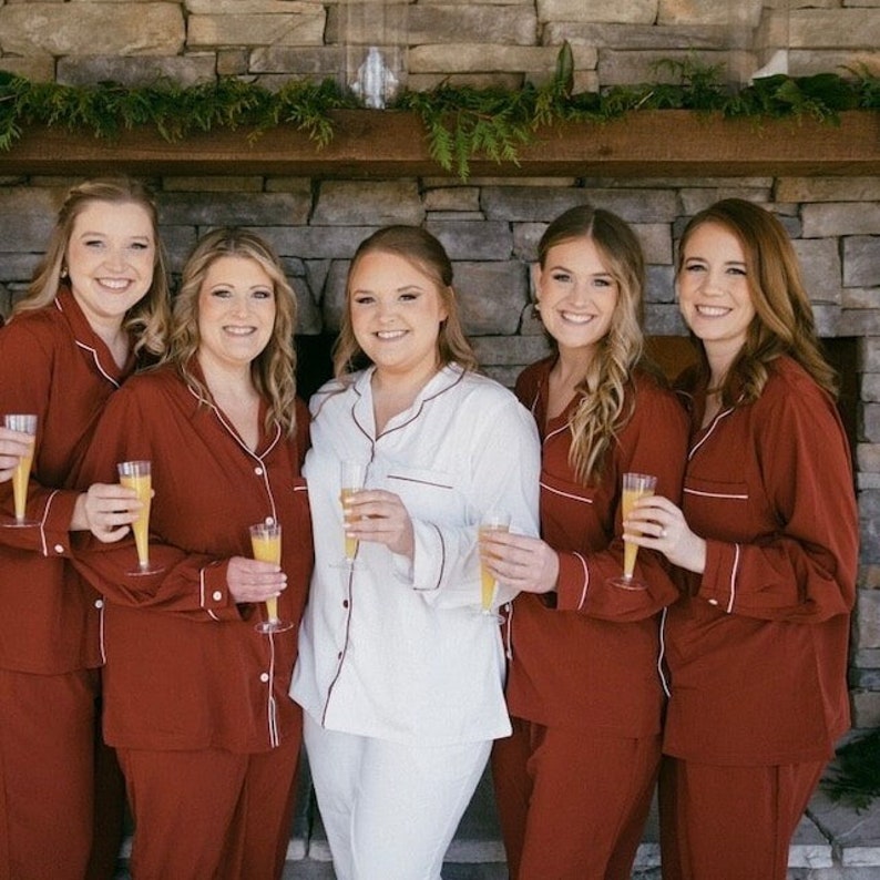 May include: Six women in matching red pajamas with white piping, standing in front of a stone fireplace, holding glasses of champagne.