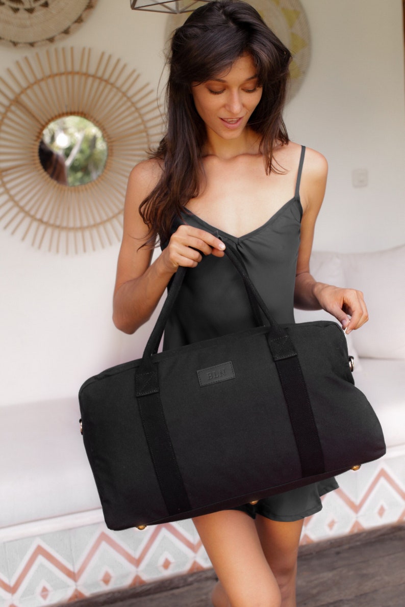 Puede incluir: Una bolsa de viaje de lona negra con un asa de cuero negro y un logotipo de metal dorado que dice "BLNK".