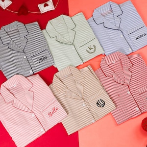 Op de afbeelding: Zes pyjamashirts met knoopsluiting, lange mouwen en kragen, voorzien van verticale strepen in grijs, lichtgroen, lichtblauw, roze, beige en rood. Elk shirt heeft een borstzak met gepersonaliseerd borduurwerk: Katie, K, JESSICA, Stella, MTS en DPB.