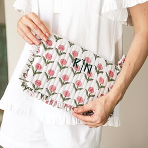 May include: A white quilted clutch bag featuring a floral design of pink flowers and green leaves. The bag has a ruffled edge and is monogrammed with the initials "KN" in dark green. The person is wearing a white top and shorts.