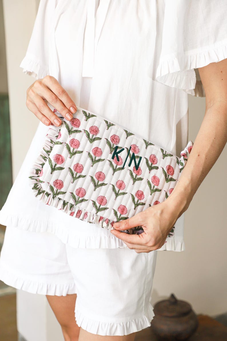 May include: A white quilted clutch bag featuring a floral design of pink flowers and green leaves. The bag has a ruffled edge and is monogrammed with the initials "KN" in dark green. The person is wearing a white top and shorts.