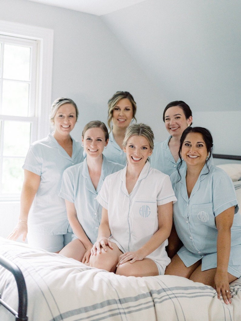 May include: Six women in matching light blue pajamas with white trim and embroidered initials. They are sitting on a white and blue striped bed. The women are smiling and looking at the camera.