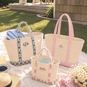Op de afbeelding: Drie canvas draagtassen in verschillende maten. De linker tas heeft een blauwe patroonrand en het monogram BJW. De rechter tas heeft een roze gingham rand en het monogram J|L. De voorste tas heeft een bloemenrand en de naam Ashlee geborduurd.