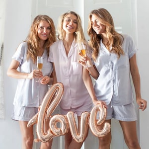 May include: Three friends wearing matching light blue and pink pyjamas hold a gold foil balloon that says "love". They are smiling and holding champagne flutes.