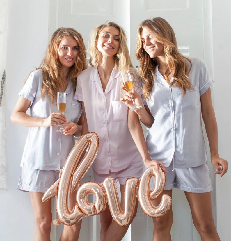 May include: Three friends wearing matching light blue and pink pyjamas hold a gold foil balloon that says "love". They are smiling and holding champagne flutes.