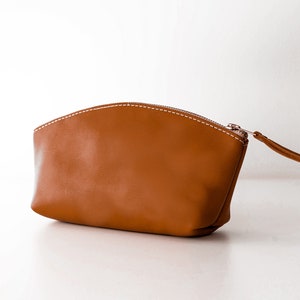 May include: A brown leather zippered pouch with white stitching. The pouch is shaped like a half-circle and has a zipper pull on the right side.