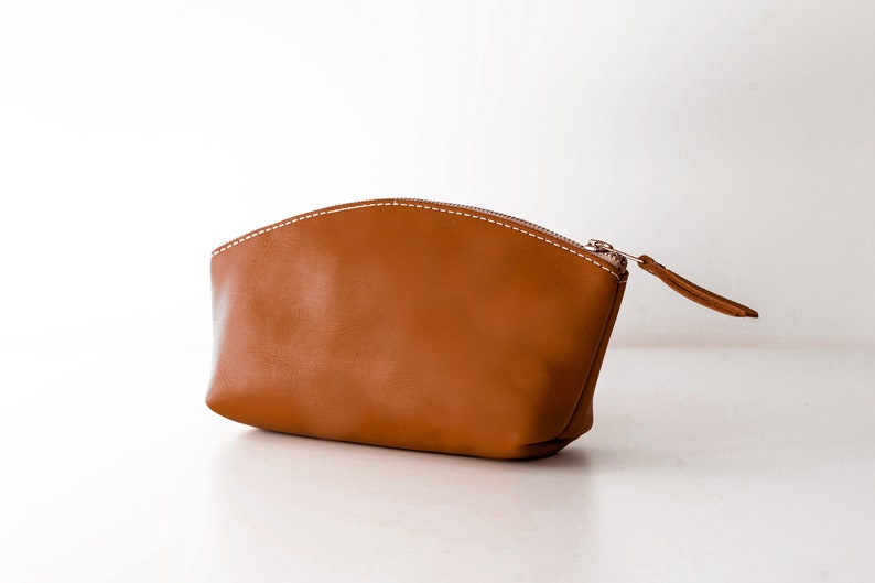 May include: A brown leather zippered pouch with white stitching. The pouch is shaped like a half-circle and has a zipper pull on the right side.