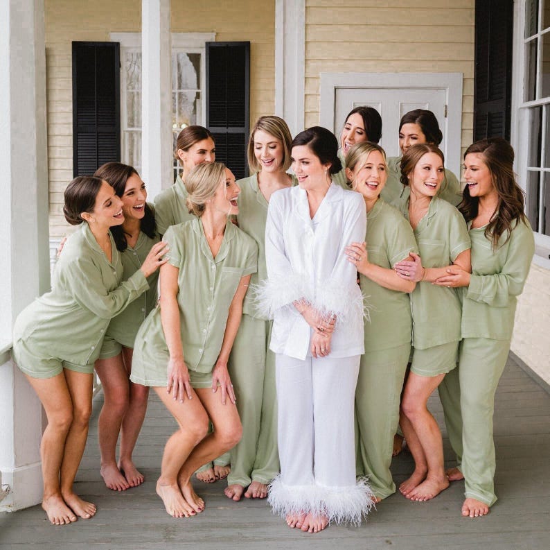 May include: A group of women wearing matching sage green pyjamas with white trim. The woman in the centre is wearing a white pyjama set with feather trim. The women are smiling and laughing together.