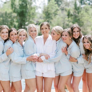 May include: A group of eight women wearing matching light blue pajamas with white trim. They are standing in front of a body of water with green trees in the background. The women are smiling and looking at the camera.