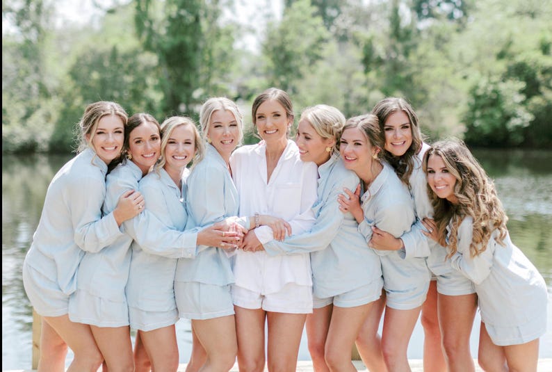 May include: A group of eight women wearing matching light blue pajamas with white trim. They are standing in front of a body of water with green trees in the background. The women are smiling and looking at the camera.