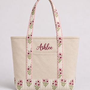 May include: A beige canvas tote bag featuring the name Ashlee embroidered in dark pink script. The bag has two handles and a bottom panel decorated with a repeating pattern of pink flowers and green leaves, accented by a pink and white gingham trim.